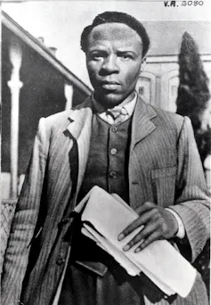 A vintage black and white photograph of a young African man, smartly dressed in a suit, who holds papers in his hand as he stands looking straight ahead in front of old buildings.