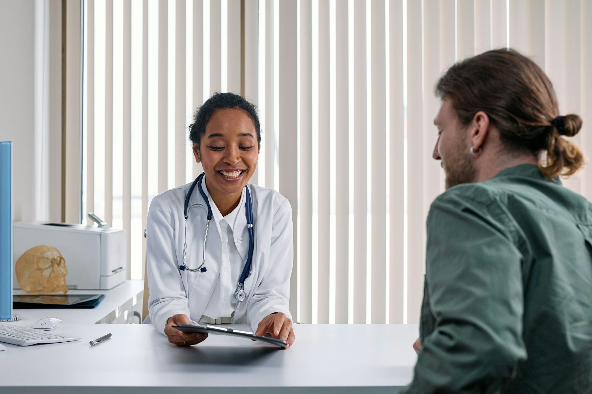 Doctor in white coat on one side of the desk similing and looking at patient, a man in a green shirt.