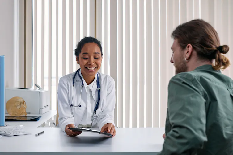 Doctor in white coat on one side of the desk similing and looking at patient, a man in a green shirt.