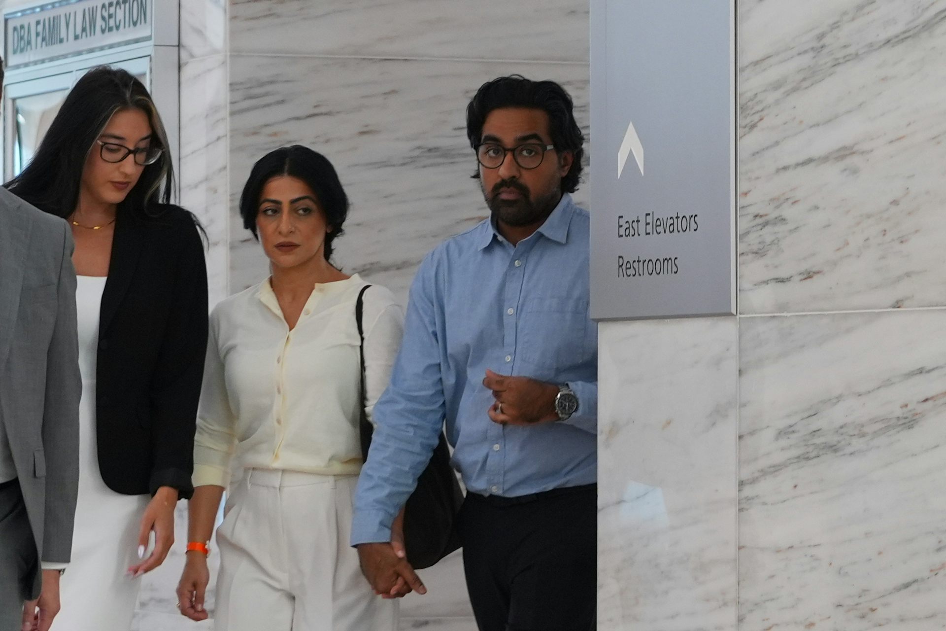 Two women and a man walk through a hallway, the man in a blue shirt holding hands with the woman in a white outfit.