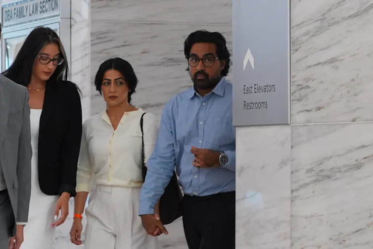 Two women and a man walk through a hallway, the man in a blue shirt holding hands with the woman in a white outfit.