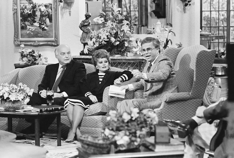 A stage adorned with flowers featuring a man wearing glasses sitting on a sofa chair next to a woman and a bald man seated on a sofa.