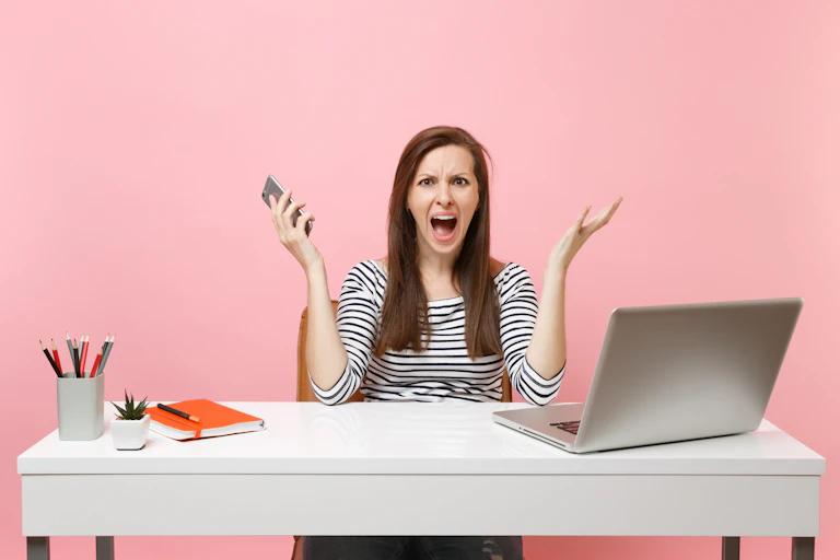 Woman looks anguished at desk with laptop and phone.