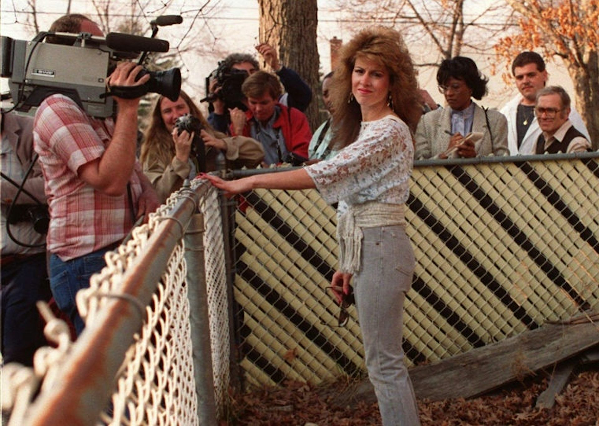 Young woman with long brown, curly hair stands against a chain link fence before a gaggle of reporters and cameramen on the other side.
