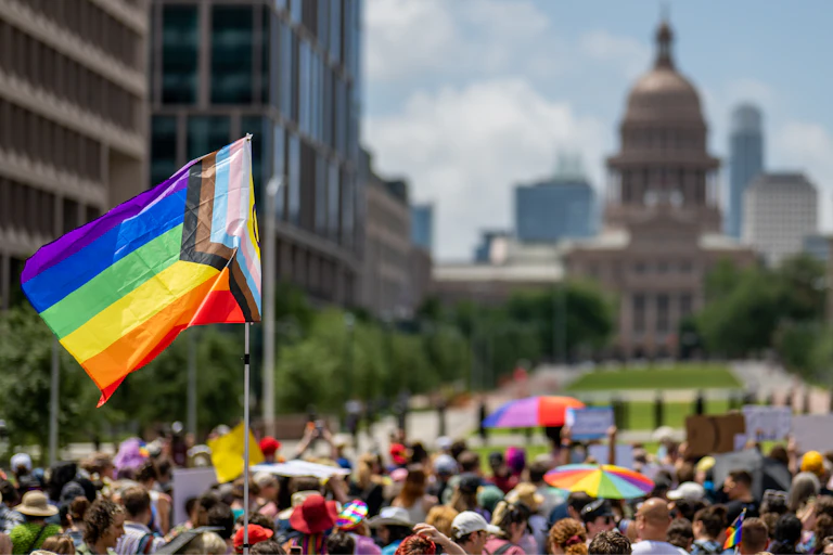 A Pride flag is held above a large crowd with a domed building in the background.