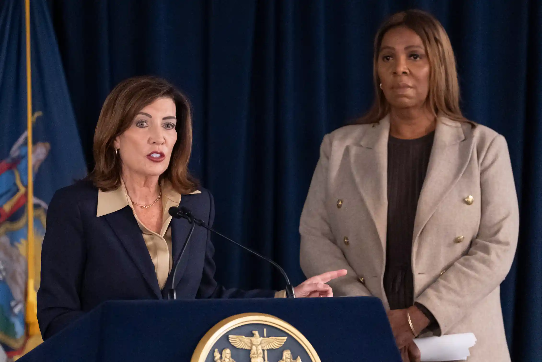 Two women look solemn as they stand behind a lectern. They are dressed in suits.