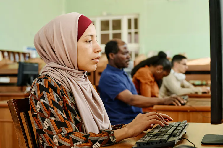 A woman wearing a head scarf sits at a computer, typing on a keyboard.