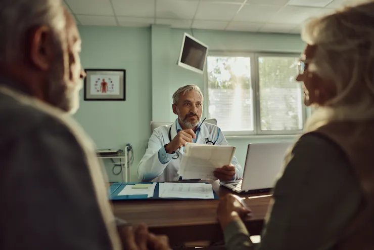 An older couple sitting in their doctor's office while he looks at some printouts.
