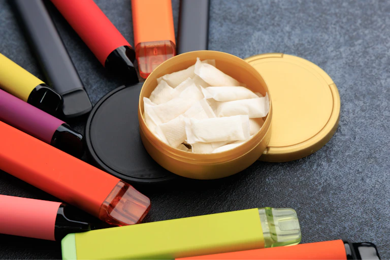 A colorful variety of vapes lying on a counter surrounding a container of nicotine pouches.