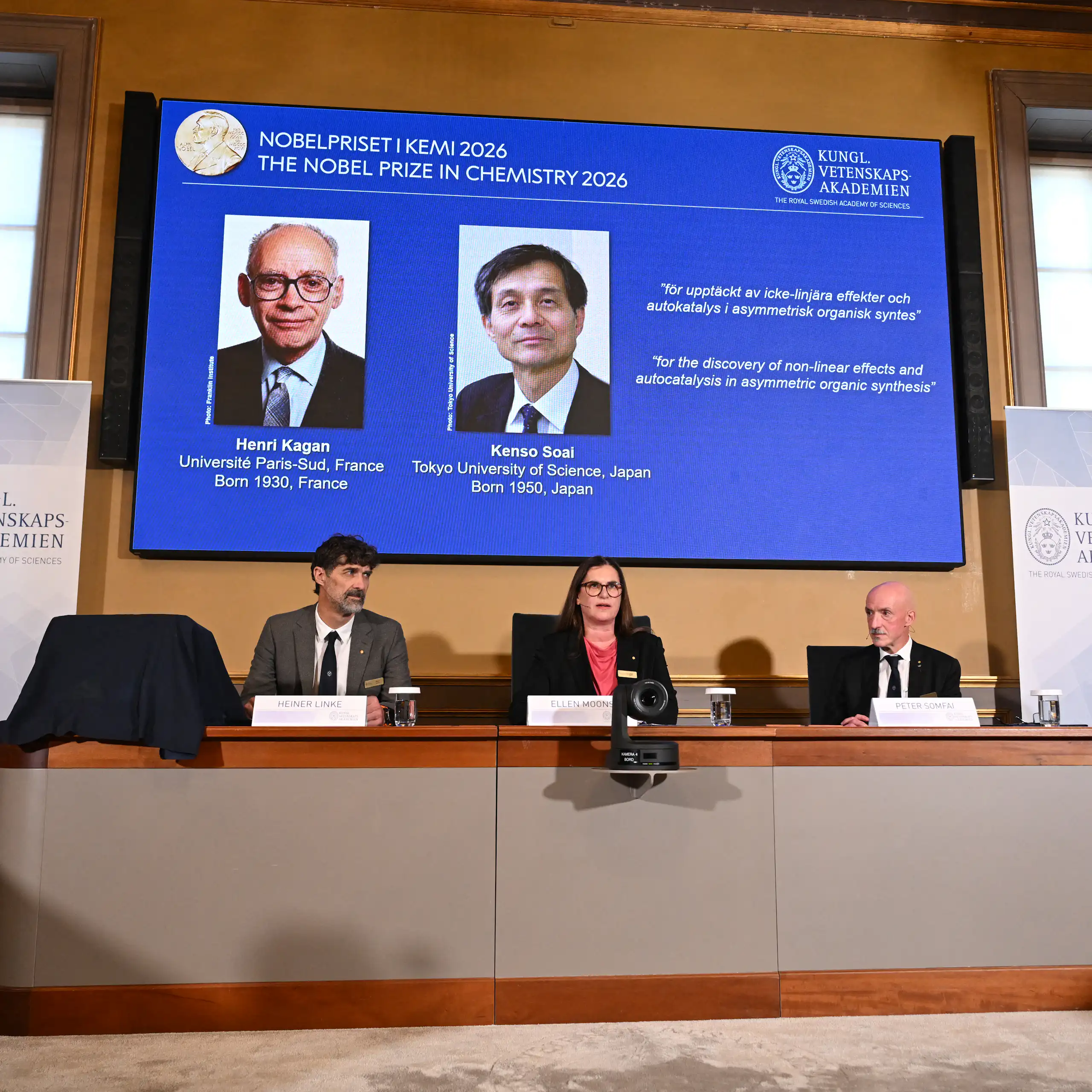 Three committee members sit at a table under a screen displaying the names and headshots of the two Nobel-winning scientists