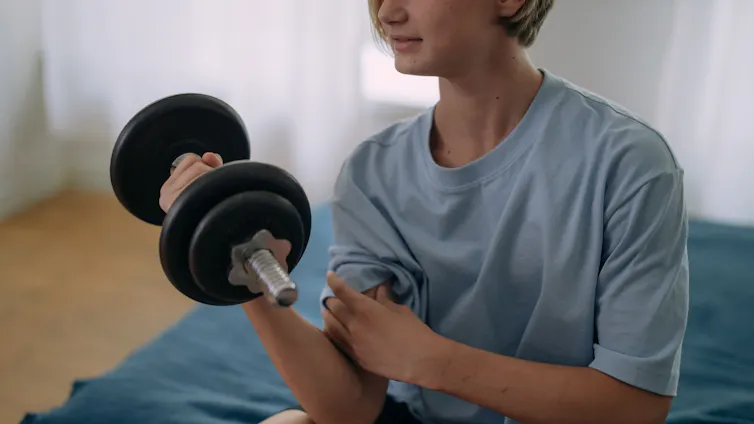 A teenage boy holds a dumbbell while sitting on a bed.