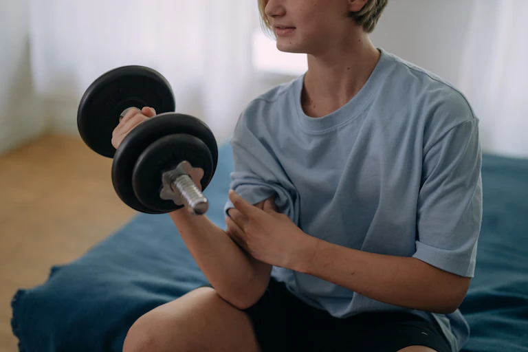 A teenage boy holds a dumbbell while sitting on a bed.