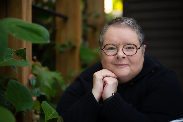 A smiling woman with short, greying hair, wearing a black jumper, by a plant, hands clasped.