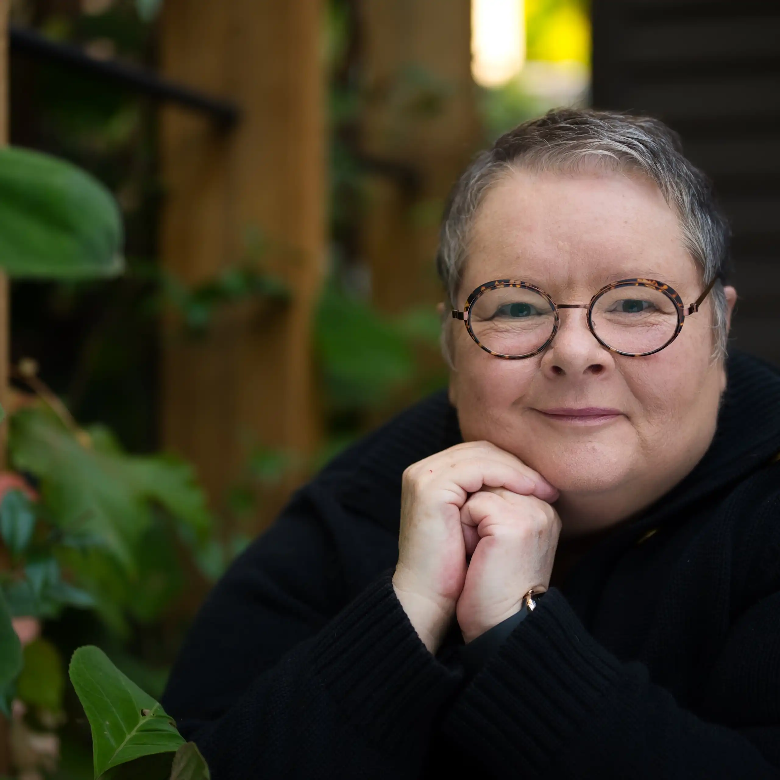 A smiling woman with short, greying hair, wearing a black jumper, by a plant, hands clasped.