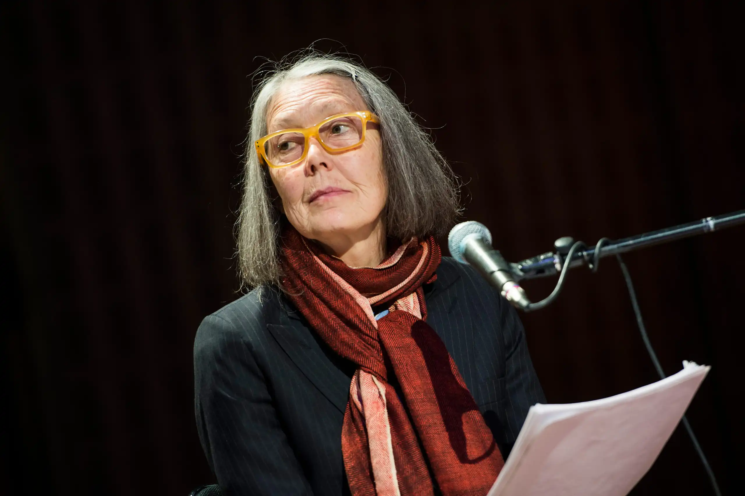 A woman at a podium in glasses and a burgundy and pink scarf gazes to the side.