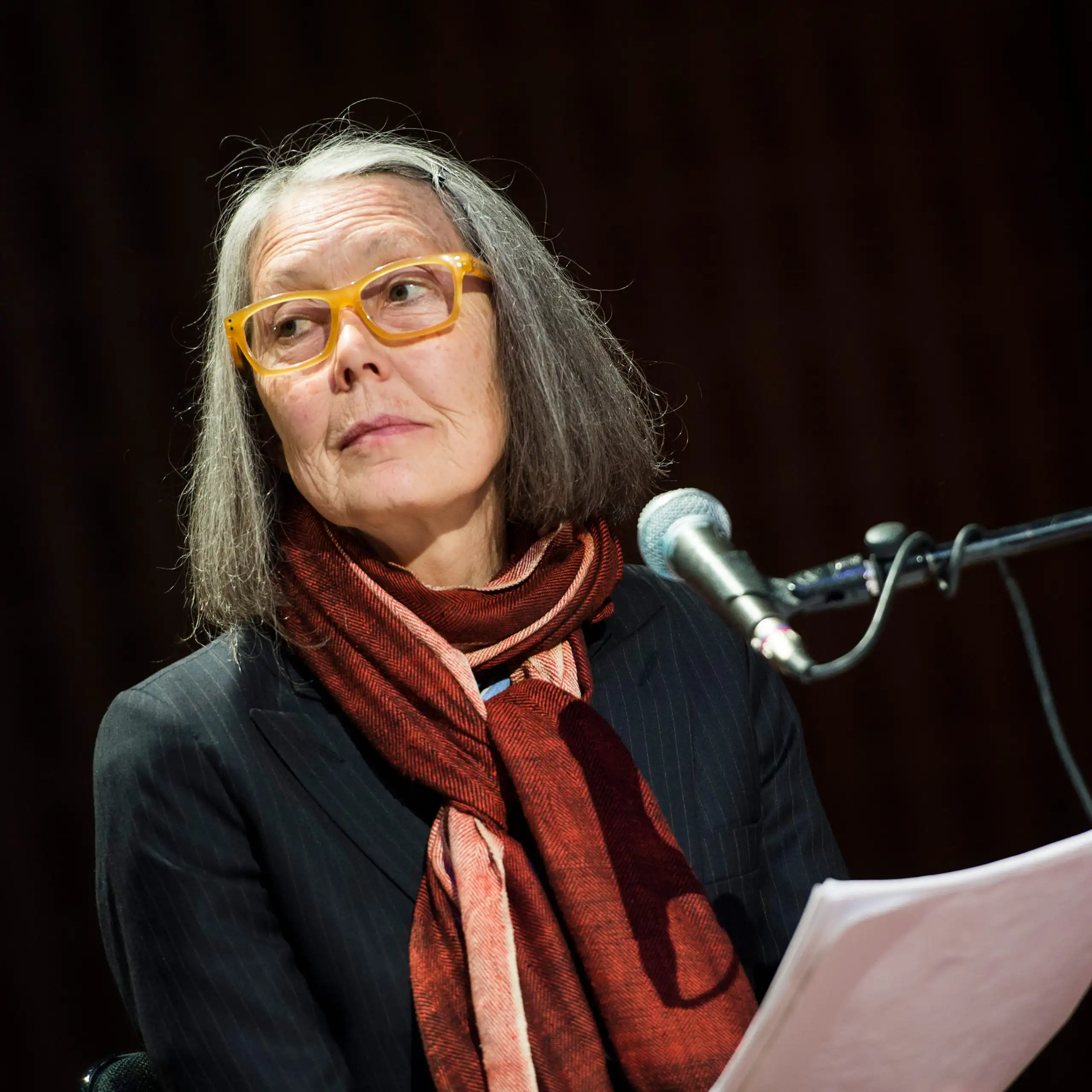 A woman at a podium in glasses and a burgundy and pink scarf gazes to the side.