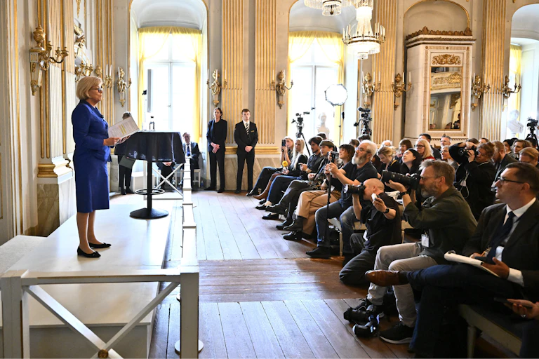 A woman at a podium in front of many photographers in a brightly lit room under a chandelier.