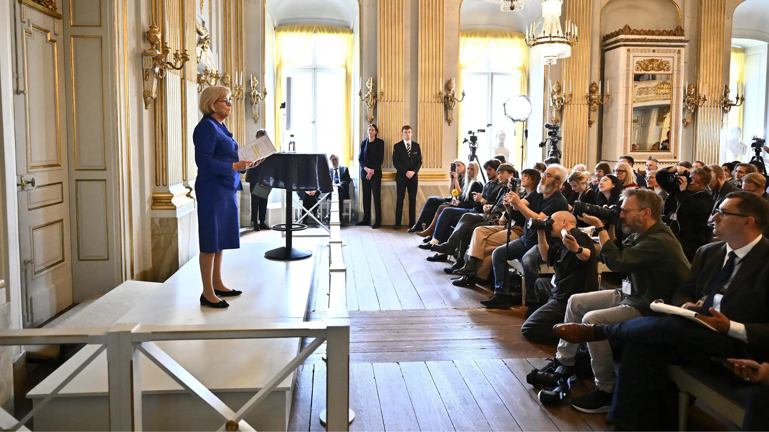 A woman at a podium in front of many photographers in a brightly lit room under a chandelier.