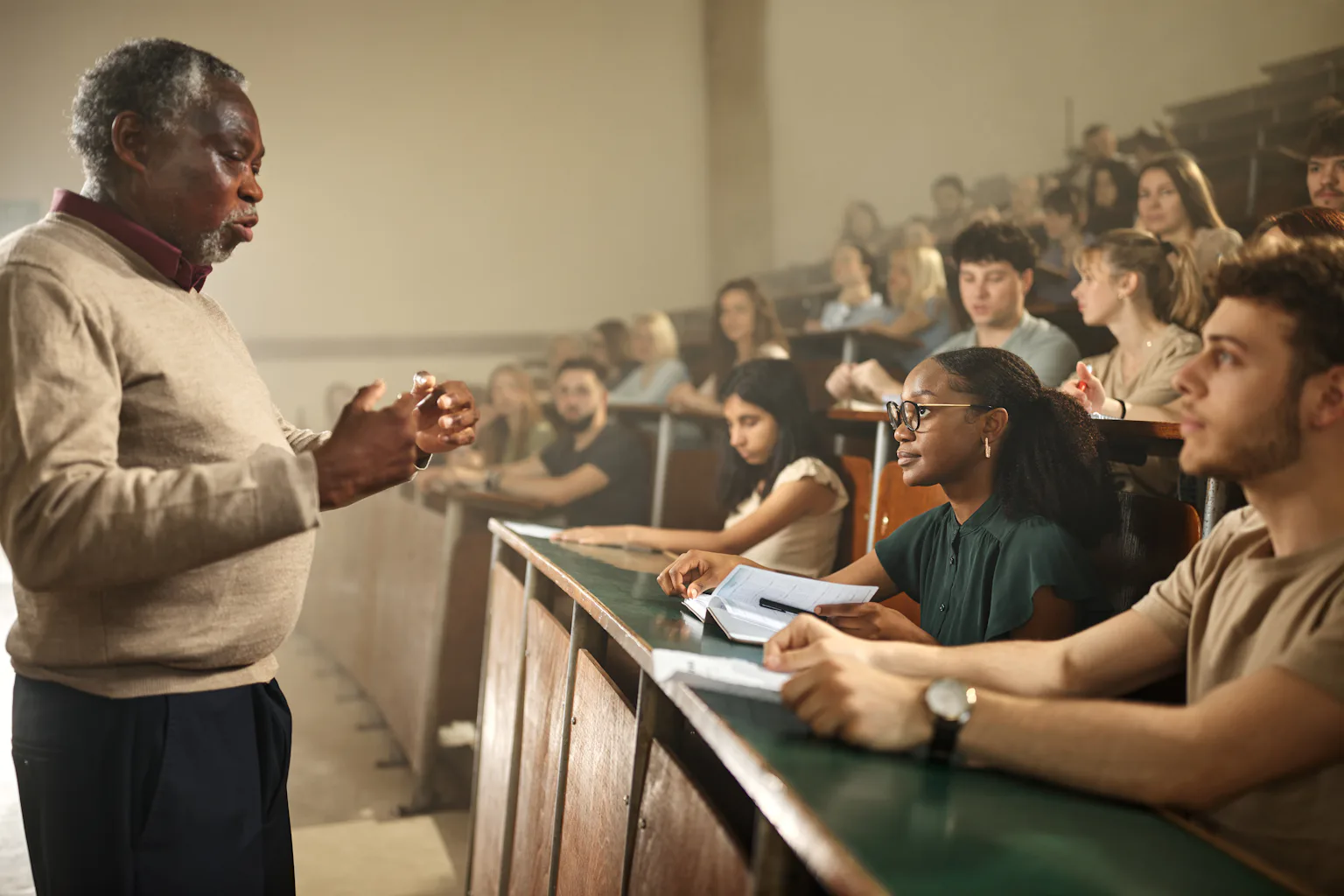 An image shows a Black older man standing with his hands extended toward a large group of college students who are seated and watching him.