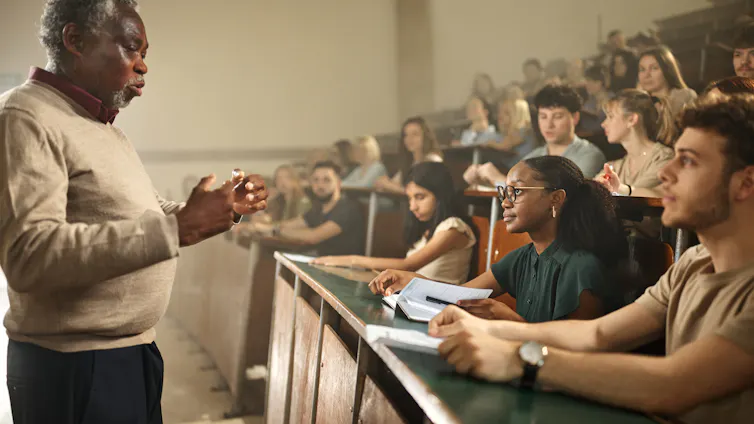 An image shows a Black older man standing with his hands extended toward a large group of college students who are seated and watching him.