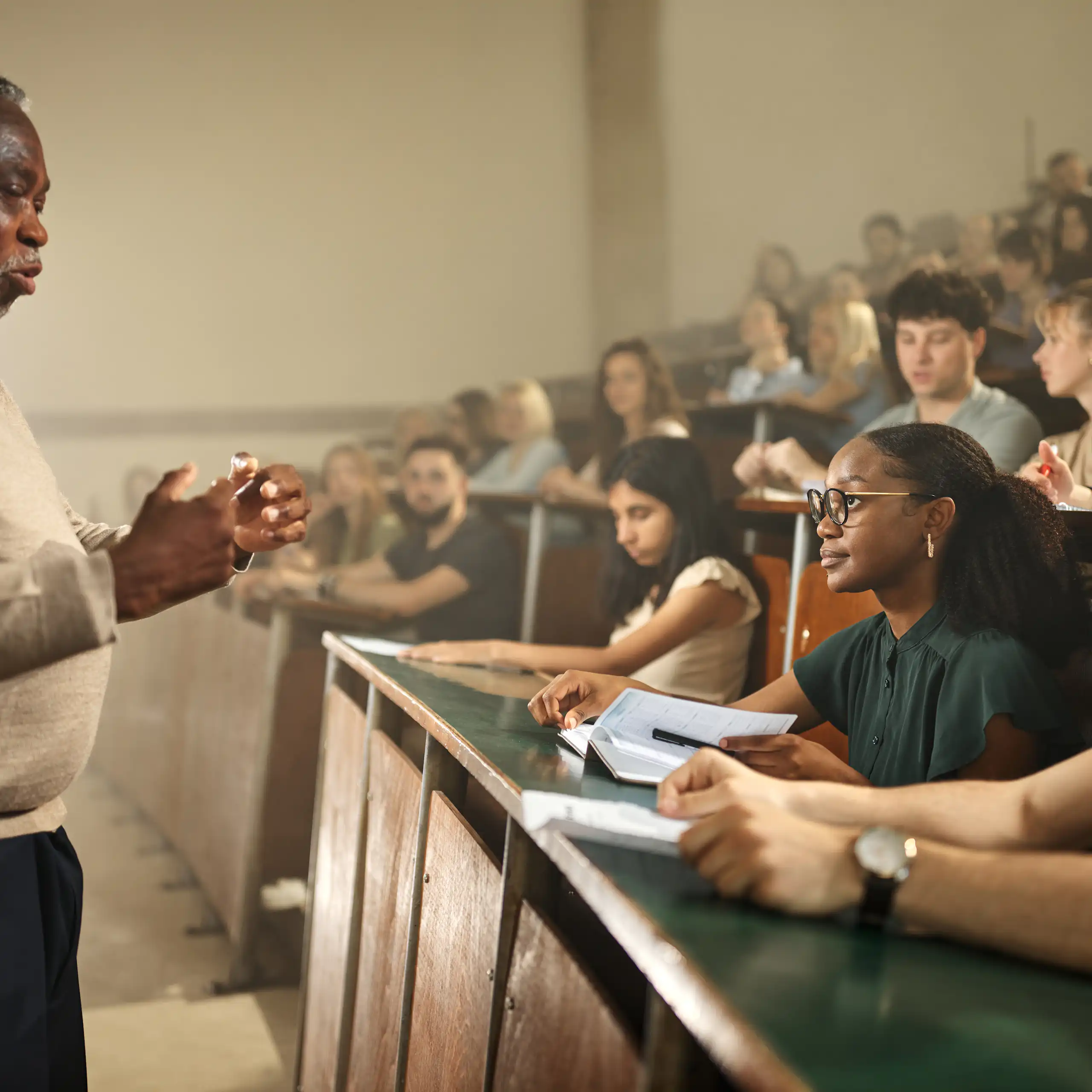 An image shows a Black older man standing with his hands extended toward a large group of college students who are seated and watching him.