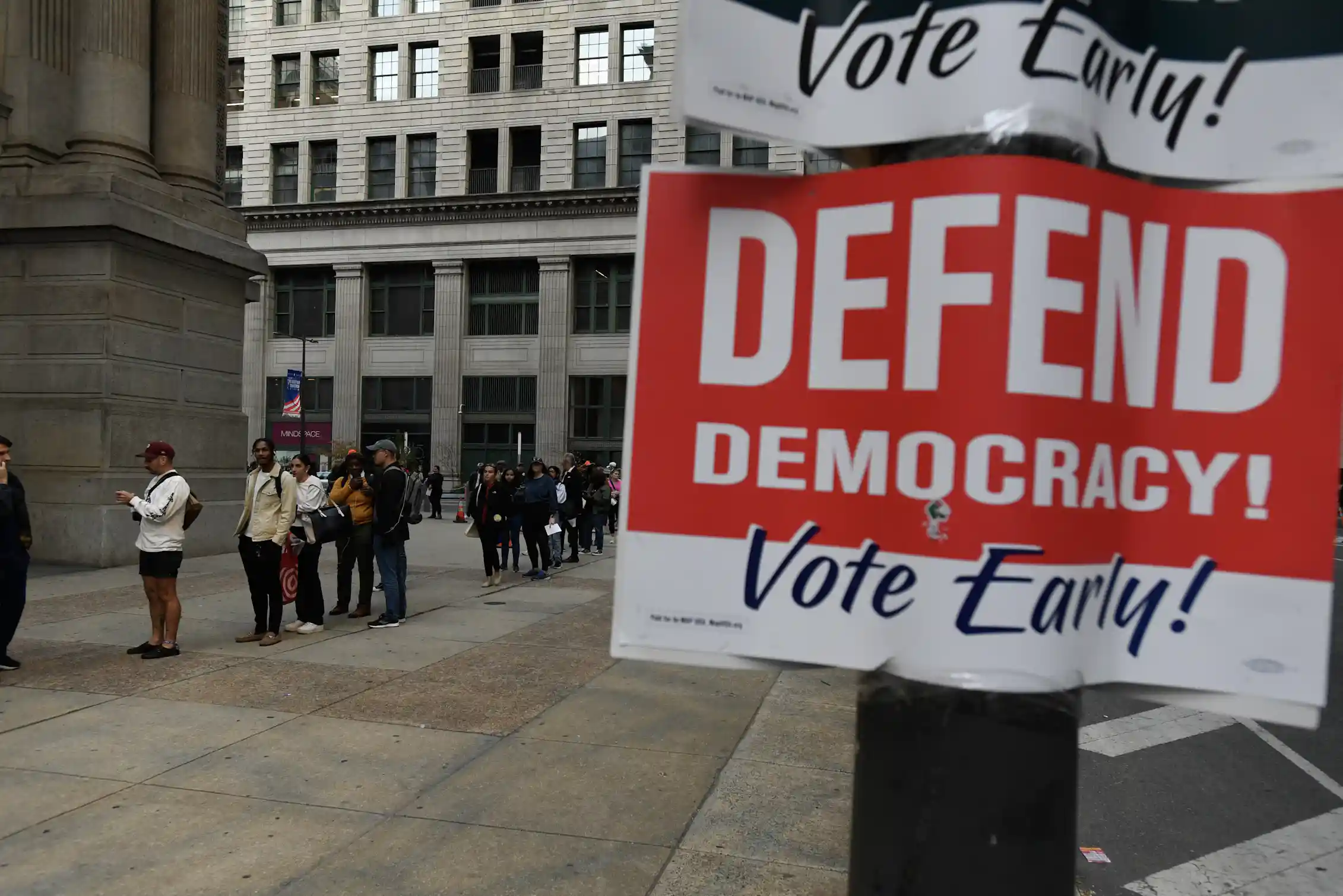 People wait in a line outside, and there's a sign that says 'Defend Democracy! Vote Early!'