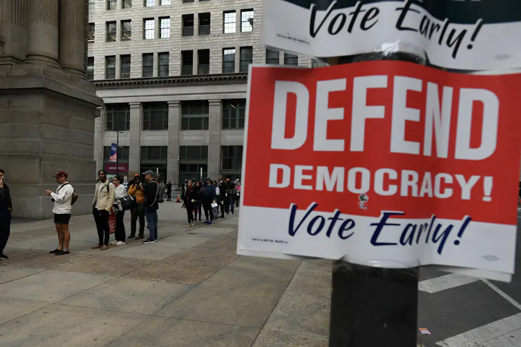 People wait in a line outside, and there's a sign that says 'Defend Democracy! Vote Early!'