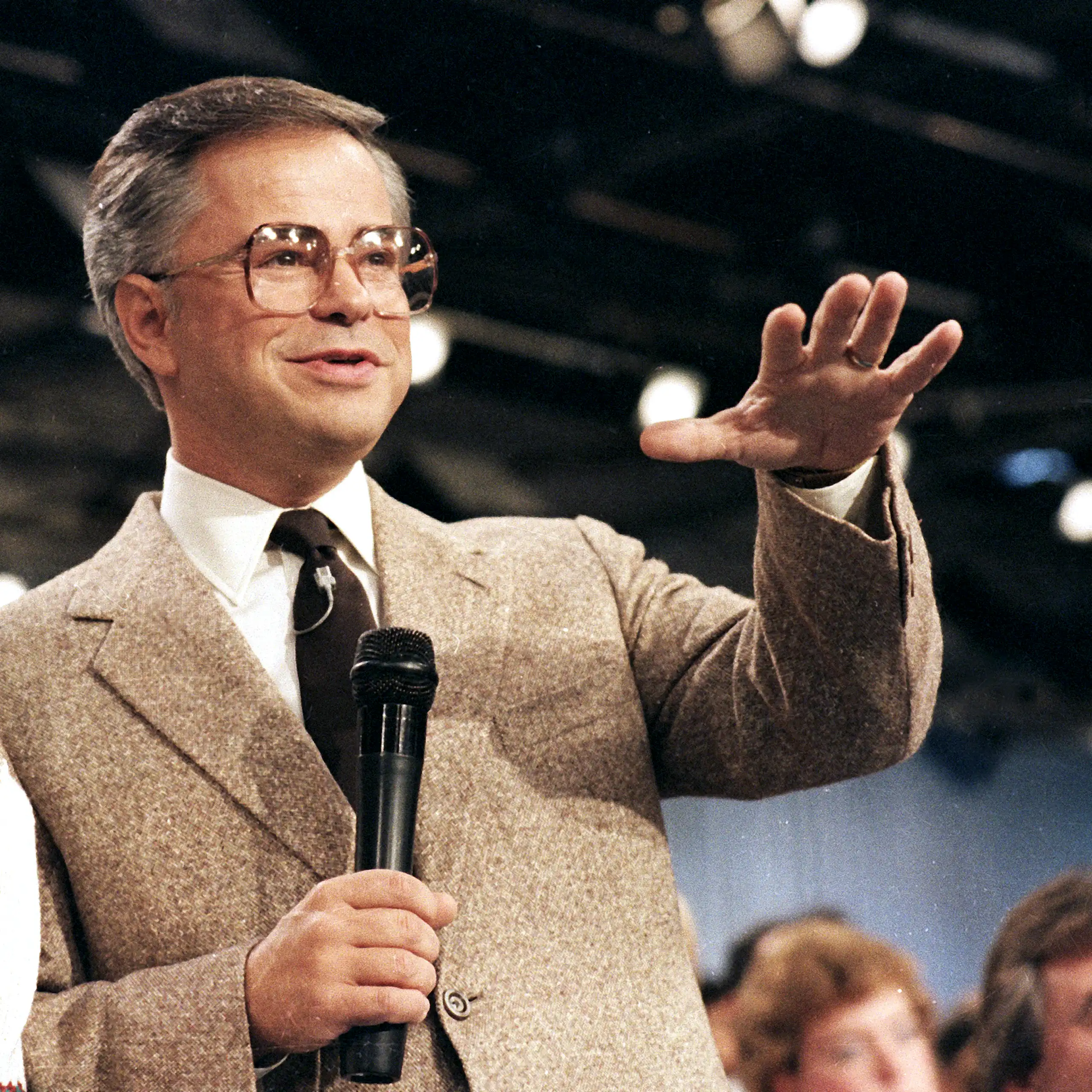 Middle-aged man wearing a suit and wide-framed eyeglasses gestures and grins while holding a microphone.