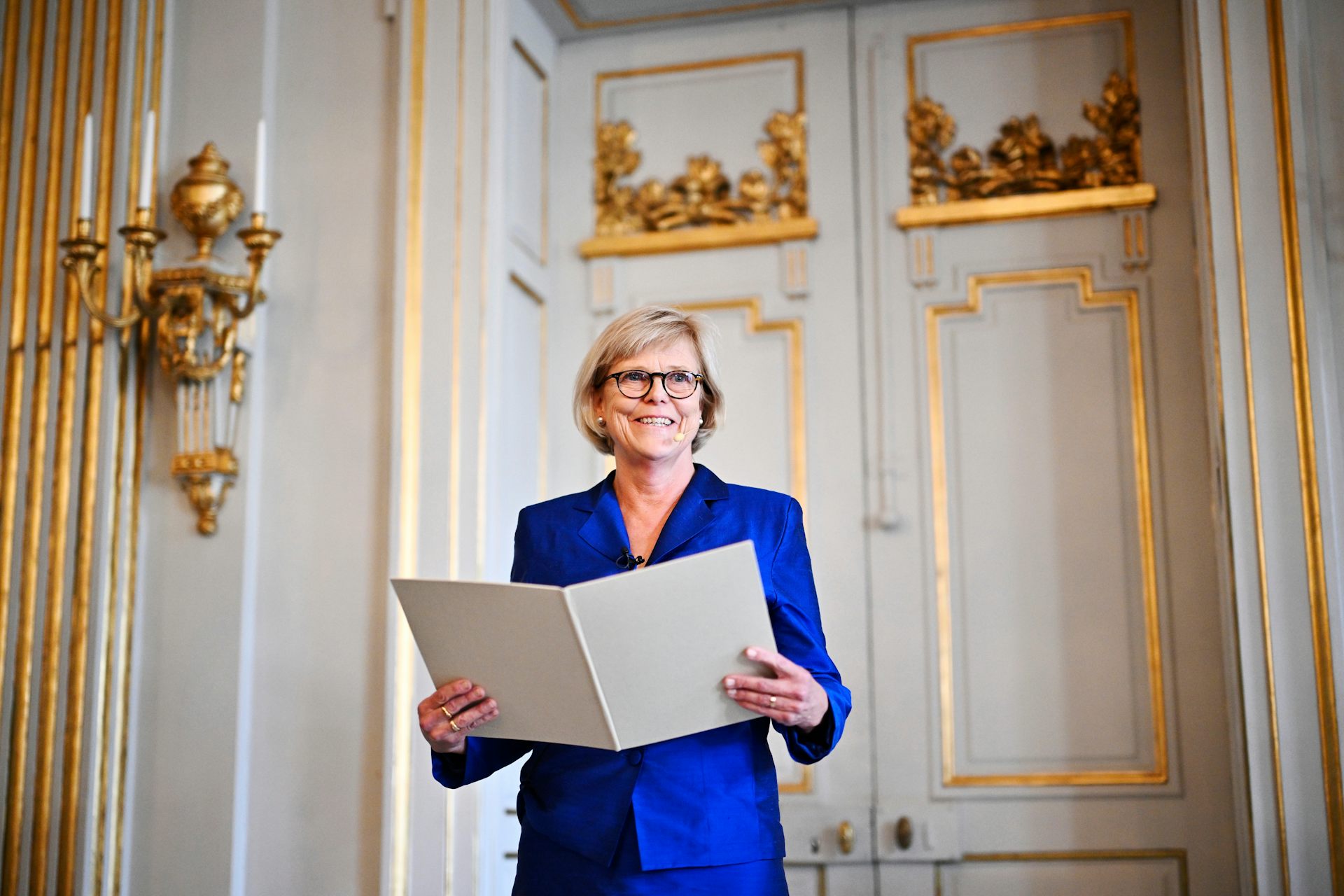 A woman in a blue dress holding a formal book in front of an ornately decorated white and gold wall.