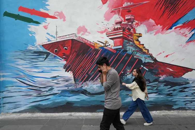 Two people walk past a mural depicting a military ship in conflict.