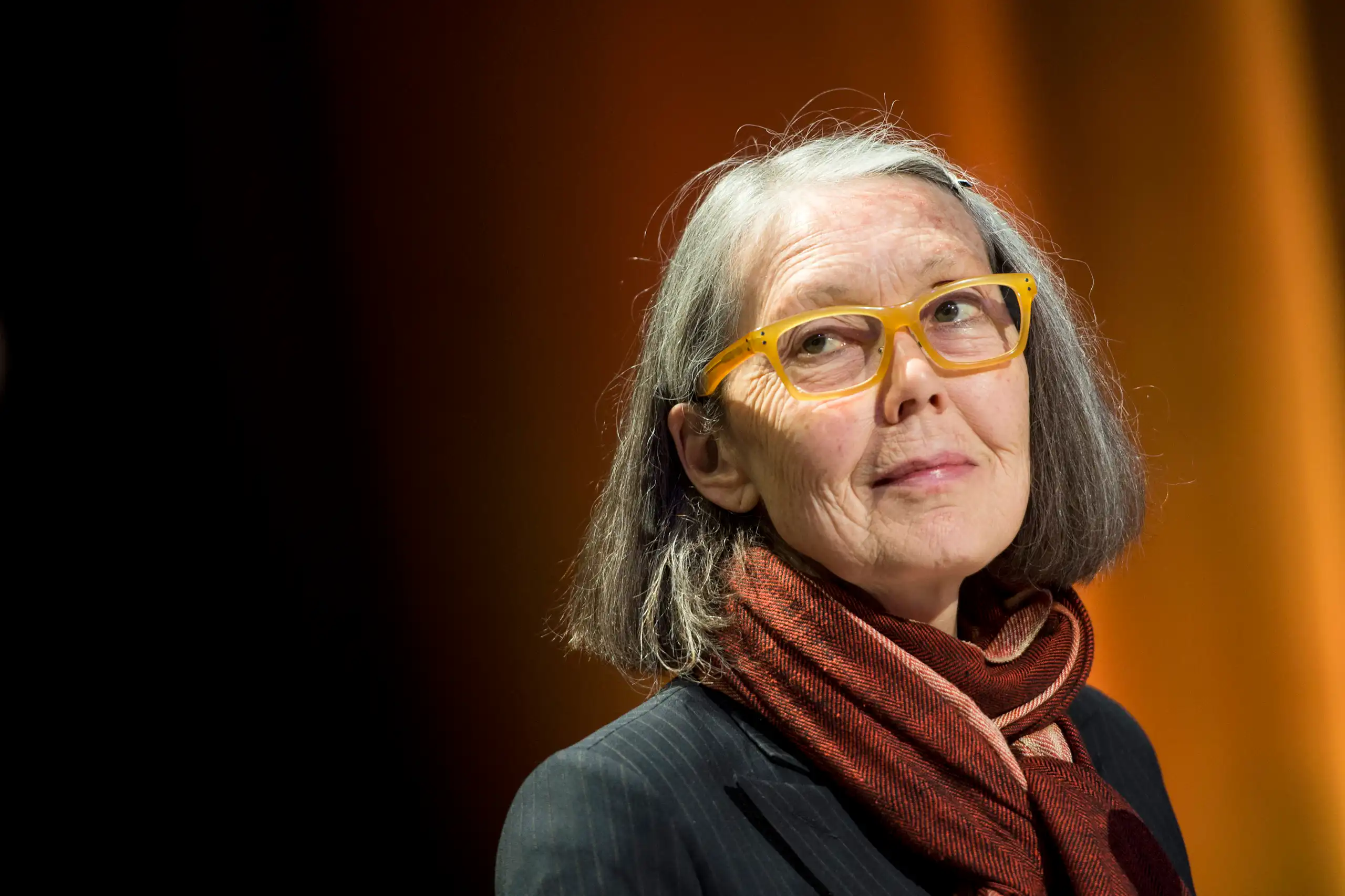 Anne Carson, a grey-haired woman with glasses and a maroon neck scarf, looks to the side, in front of a curtain