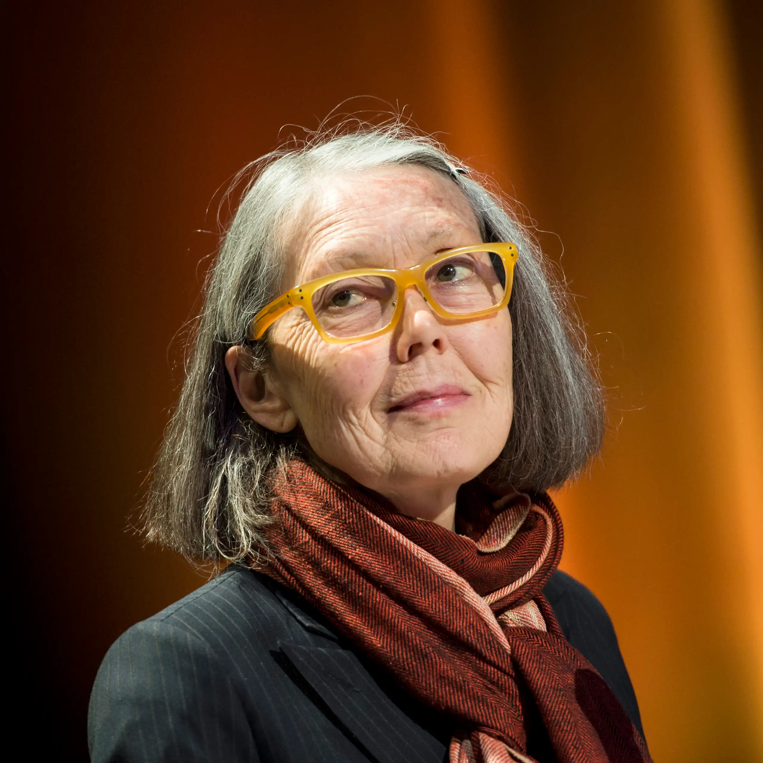 Anne Carson, a grey-haired woman with glasses and a maroon neck scarf, looks to the side, in front of a curtain