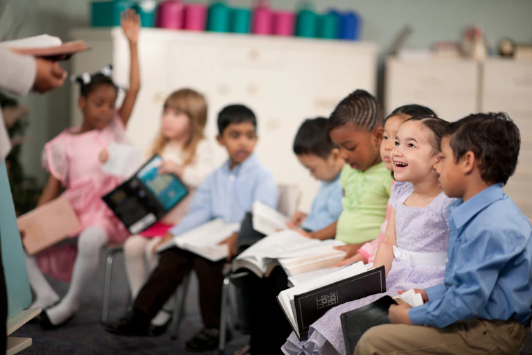 A row of young children sit in chairs and hold open books inside a classroom.