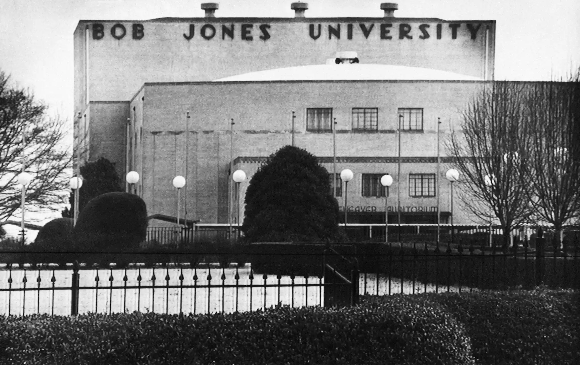 A black-and-white photo of a building, seen from behind a fence, that says 'Bob Jones University' in large letters near the roof.