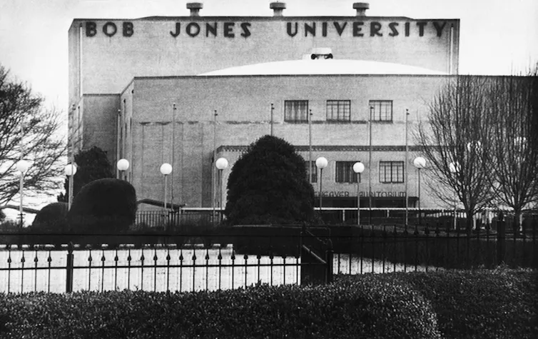 A black-and-white photo of a building, seen from behind a fence, that says 'Bob Jones University' in large letters near the roof.