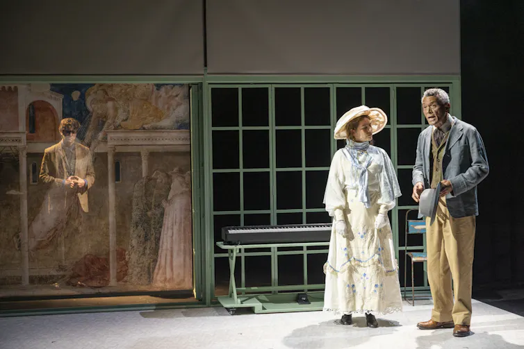 Production image: a man behind a scrim while two people talk in front of green framed windows.