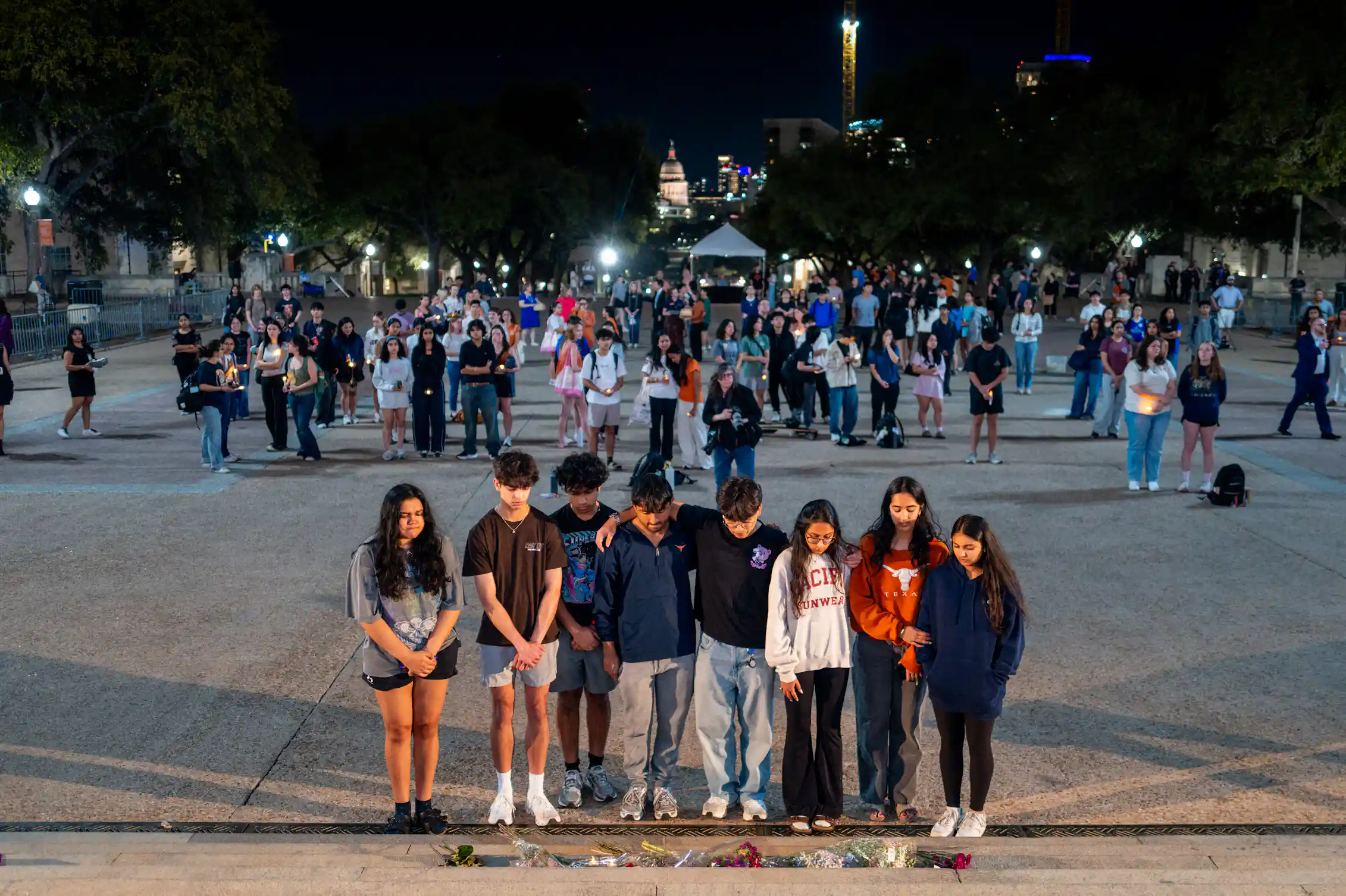 Young people stand with heads bowed behind a line of flowers on a plaza at night.
