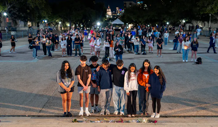 Young people stand with heads bowed behind a line of flowers on a plaza at night.