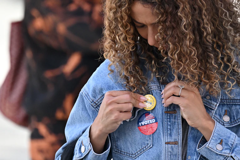 A young woman with curly hair and wearing an 'I Voted' sticker fastens a pin to her denim jacket.