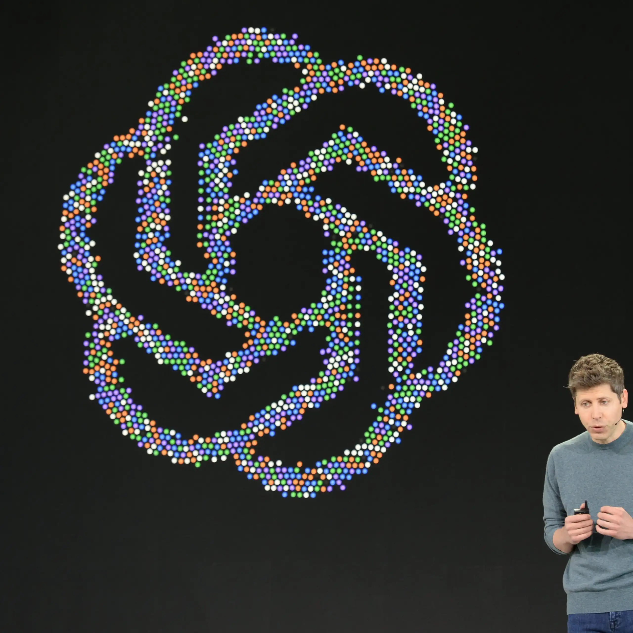 OpenAI CEO stands on stage in front of a large colourful OpenAI logo