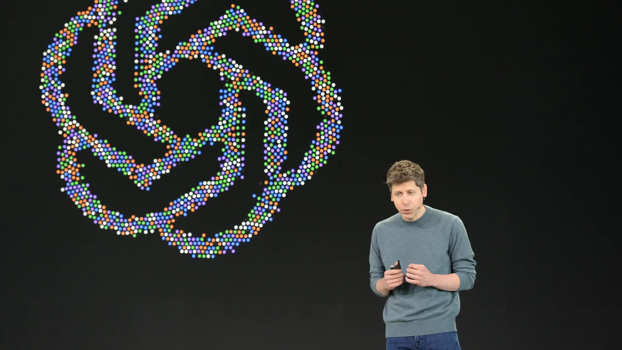 OpenAI CEO stands on stage in front of a large colourful OpenAI logo