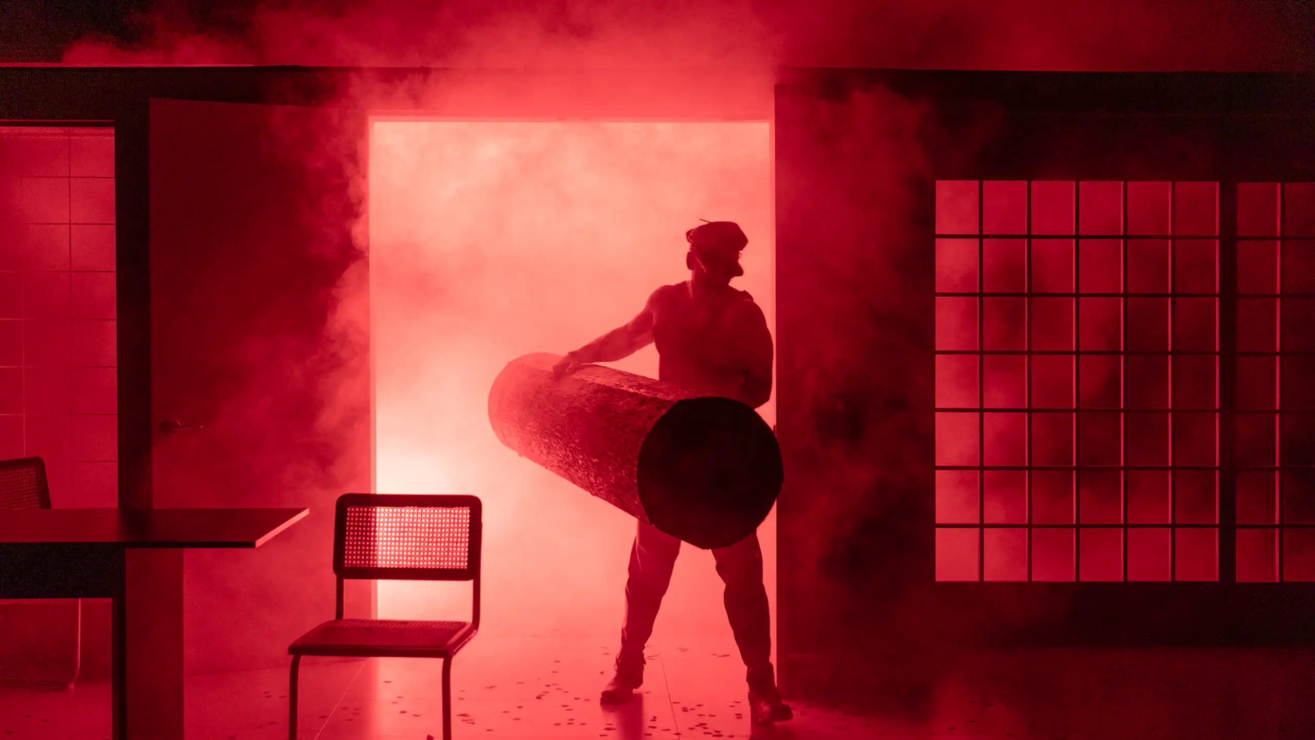 Production image: a 'leather daddy' holding a log emerges from red smoke.