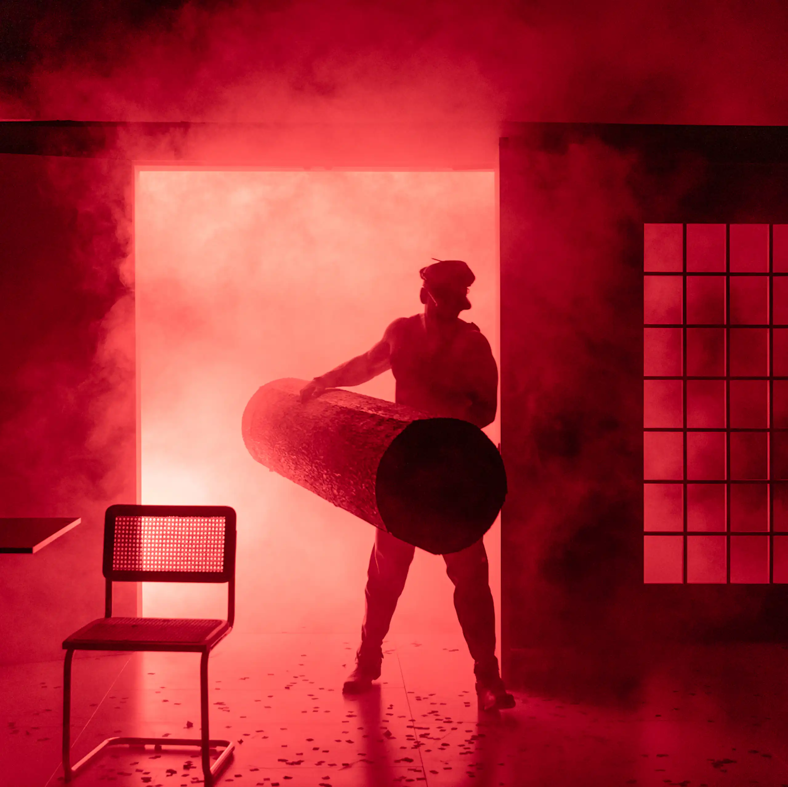 Production image: a 'leather daddy' holding a log emerges from red smoke.