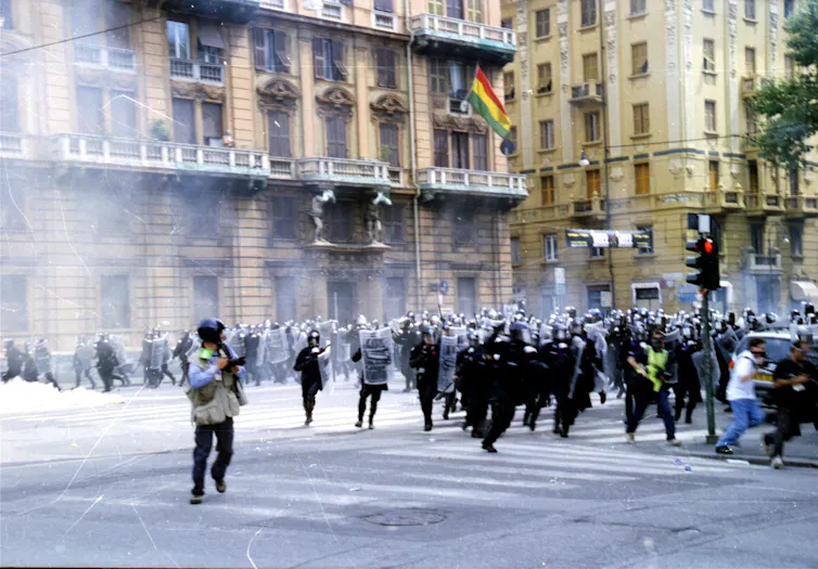 Imagen de fotógrafos de prensa corriendo delante de unos policías que cargan durante unas protestas en la calle de una ciudad.