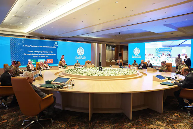 Men sit around a round table, some in Arab clothing, with the logo of the Mecca pact in the background.