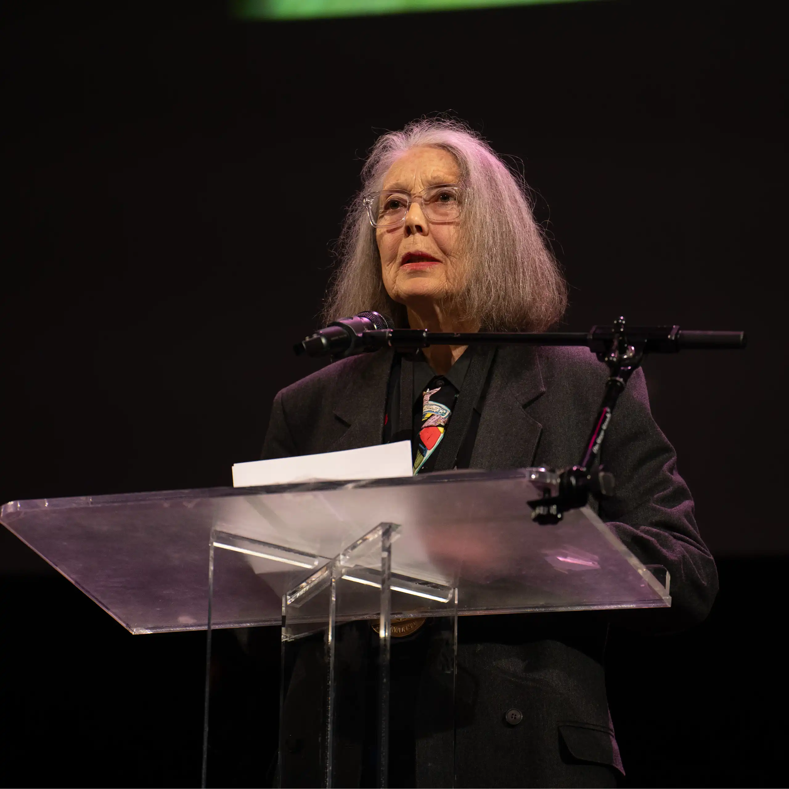 Elderly woman with straight, shoulder-length, gray hair wearing glasses and a black suit jacket stands a podium.