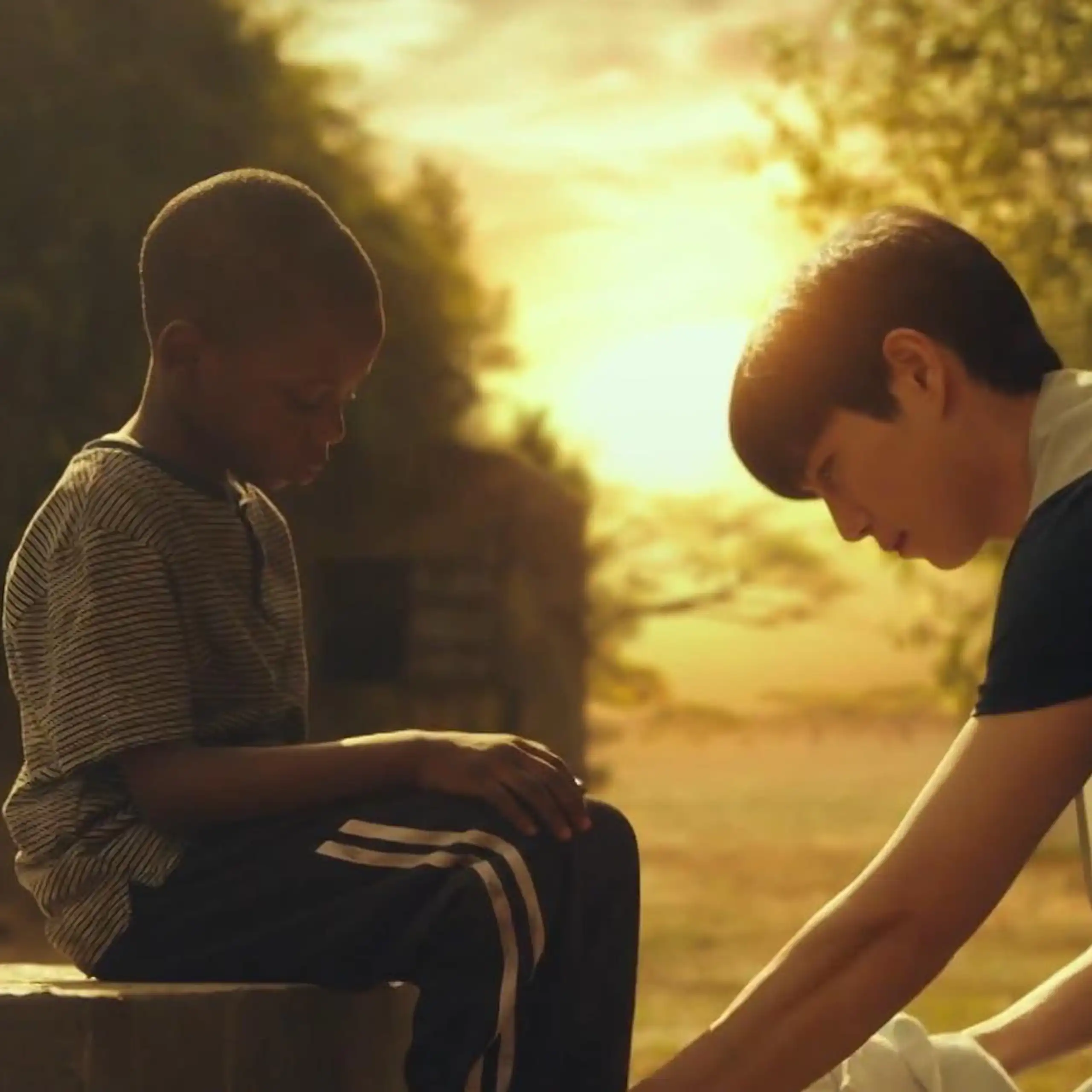 An Asian man kneels in front of an African child, the sun glowing in the background.