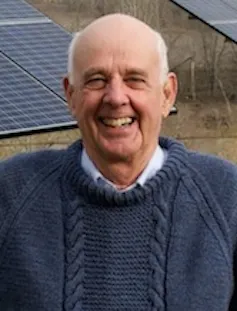 A headshot of an older balding man, who smiles in a blue sweater, in front of solar panels