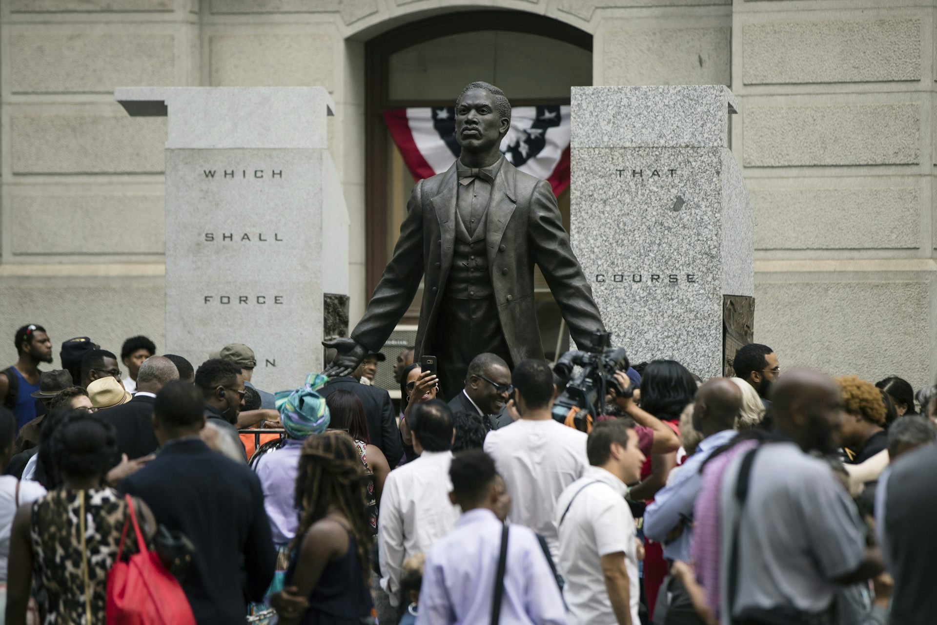 Large group of people surround a bronze statue of man
