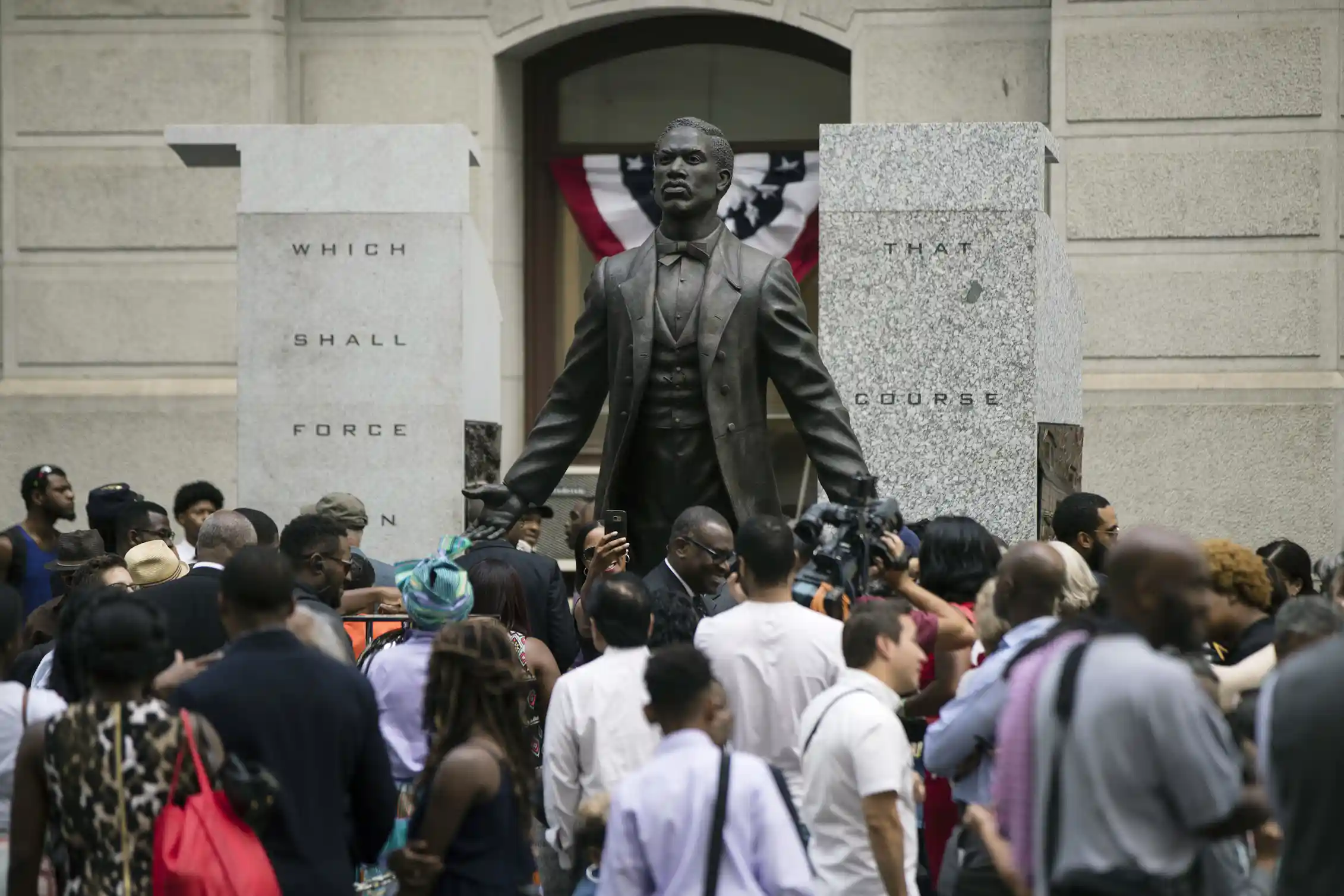 Large group of people surround a bronze statue of man